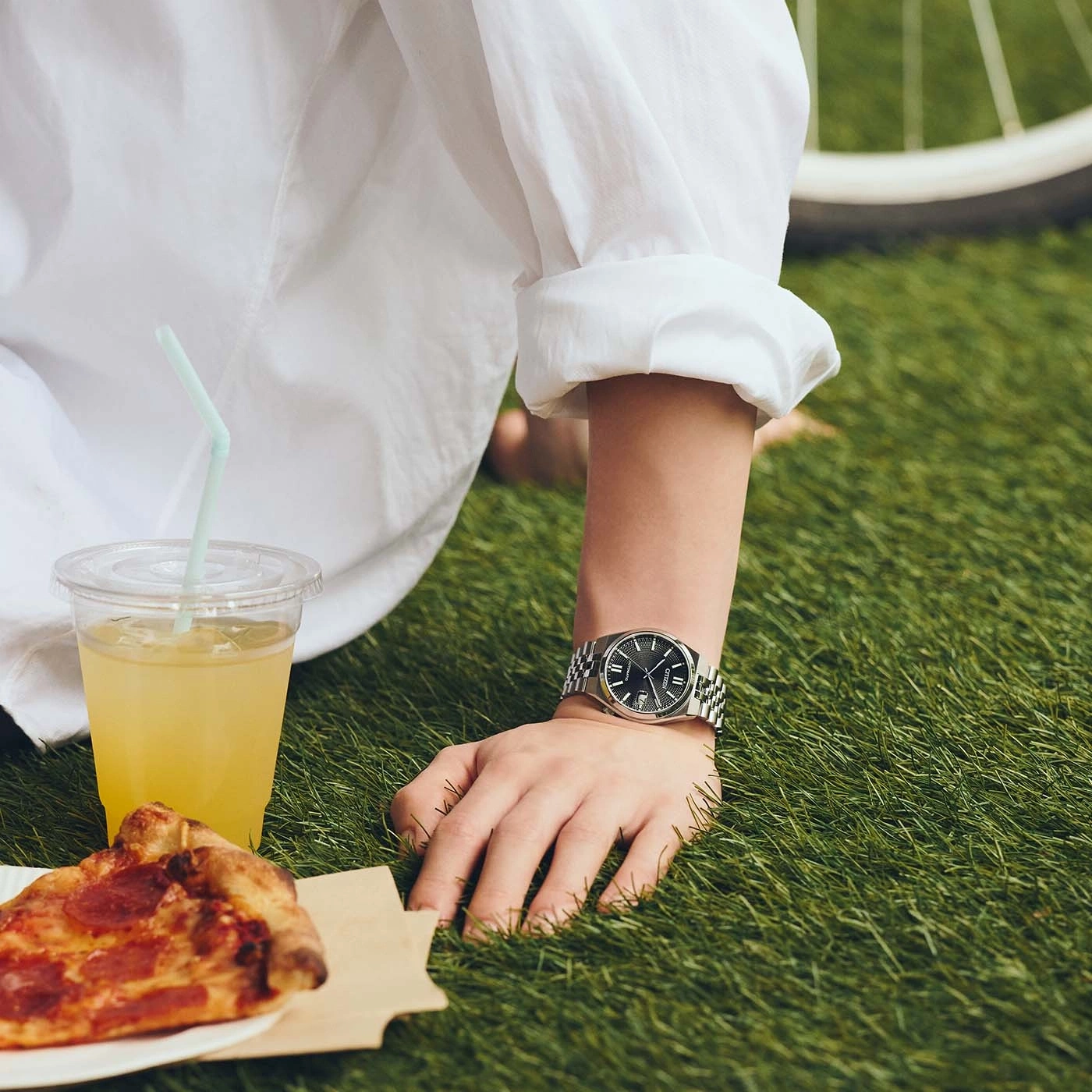 A person in a white shirt enjoys a picnic with pizza and a drink.