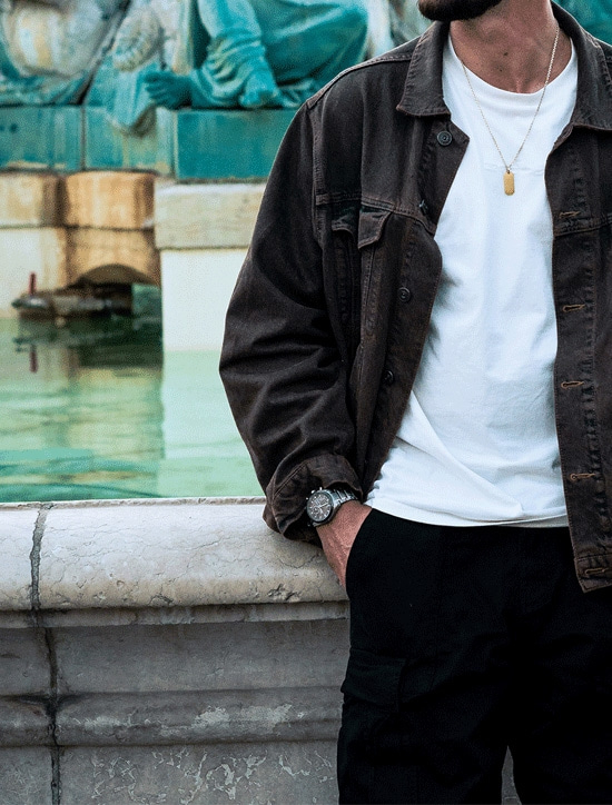 Man in brown jacket and black pants, leaning on a stone structure