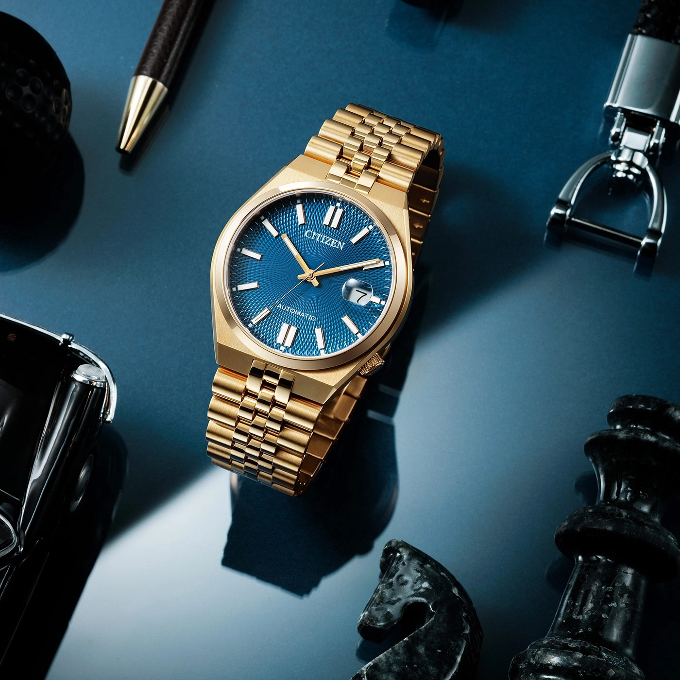 Gold Citizen watch with blue face, pen, chess pieces, and keychain on blue surface.