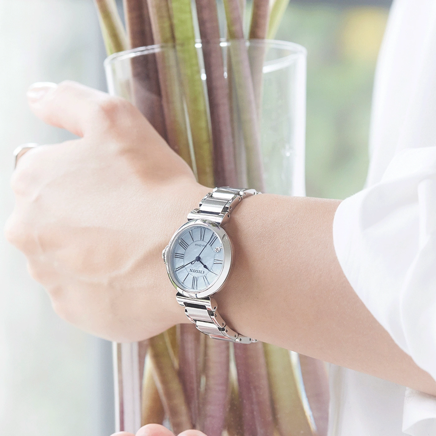 A woman's wrist with a silver watch, holding a glass vase with plants.