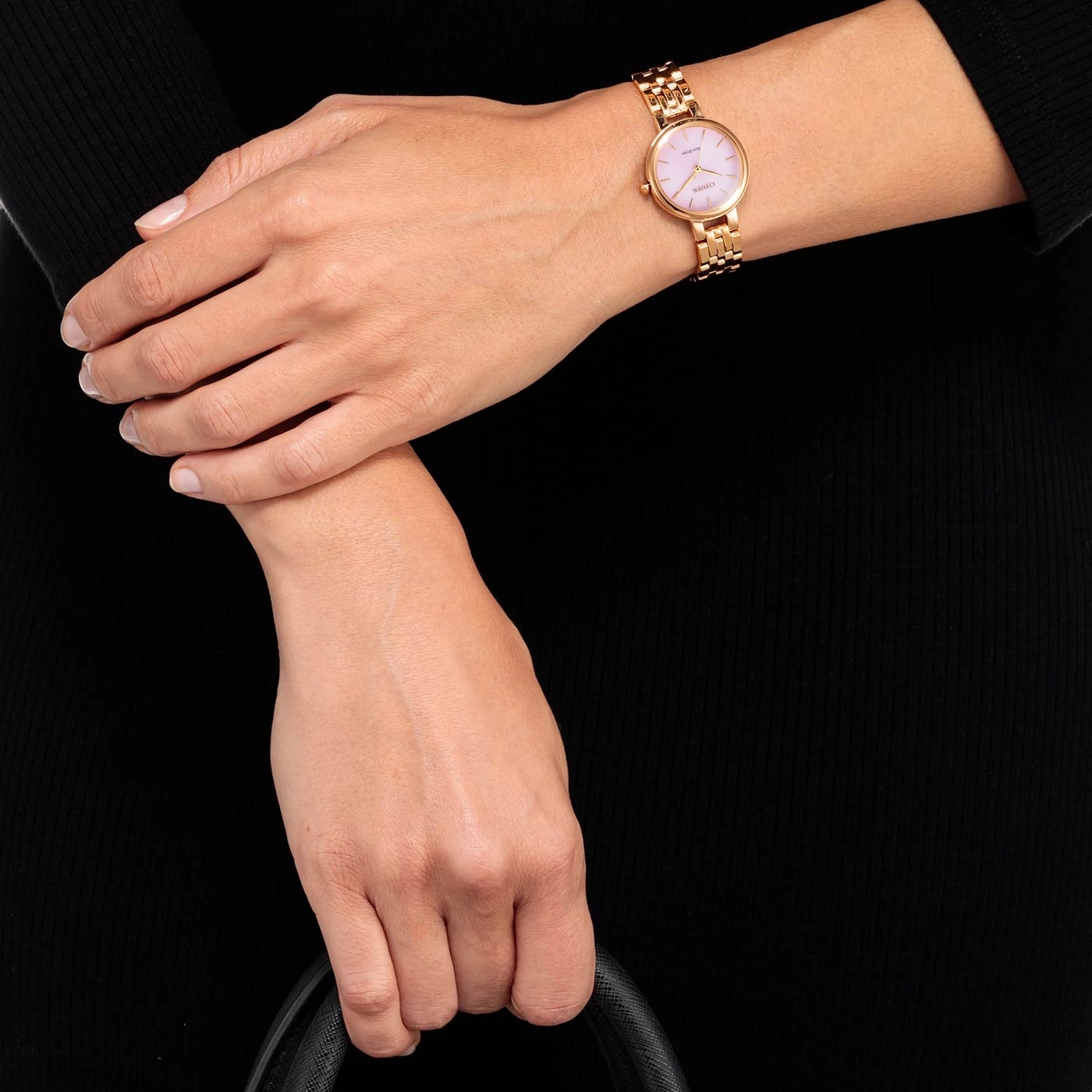 Woman wearing a gold watch with a pink face, holding a black bag.