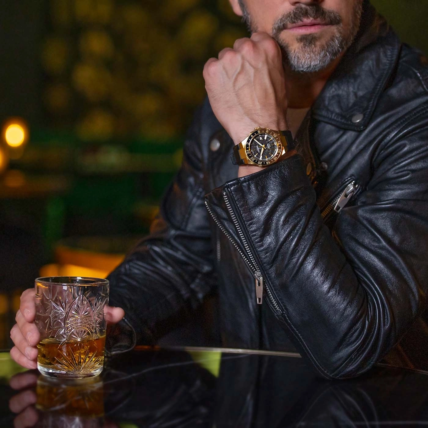 Man in leather jacket with watch holds whiskey glass.