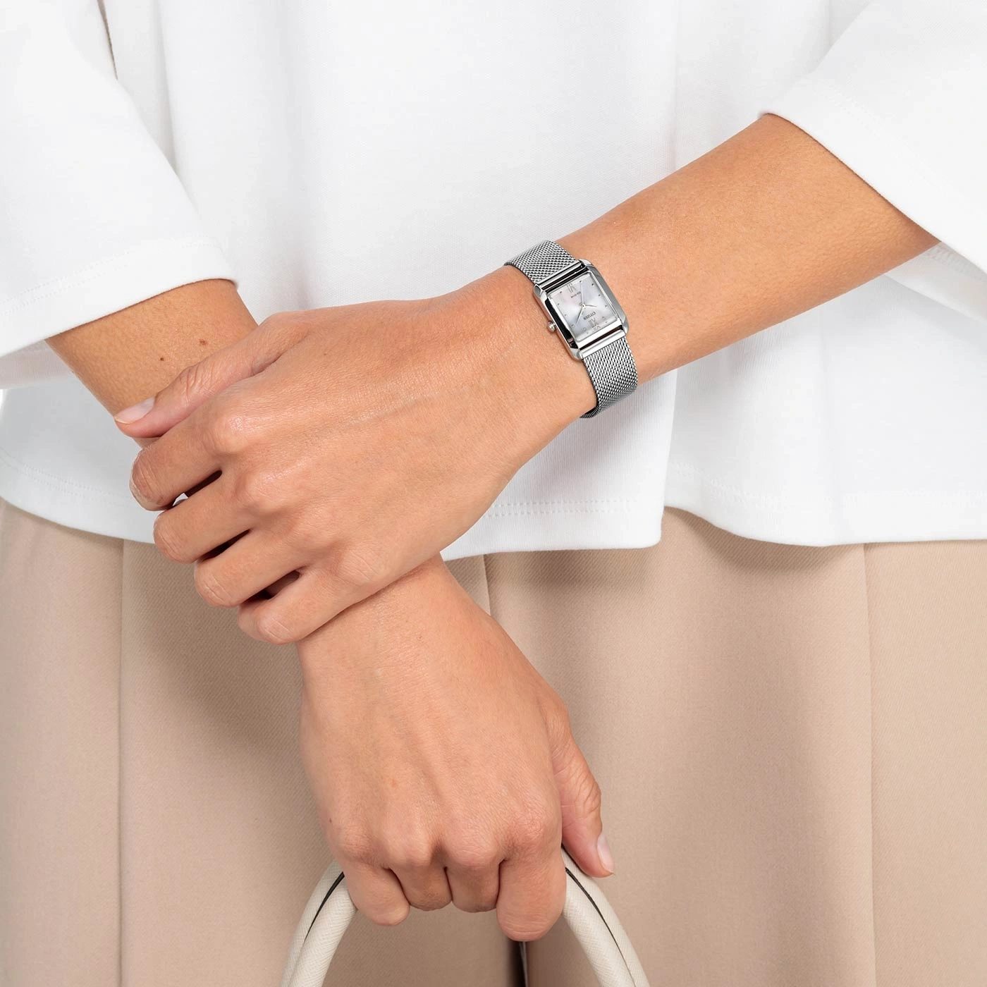 Woman wearing a silver watch, holding a white purse, beige pants and white shirt.
