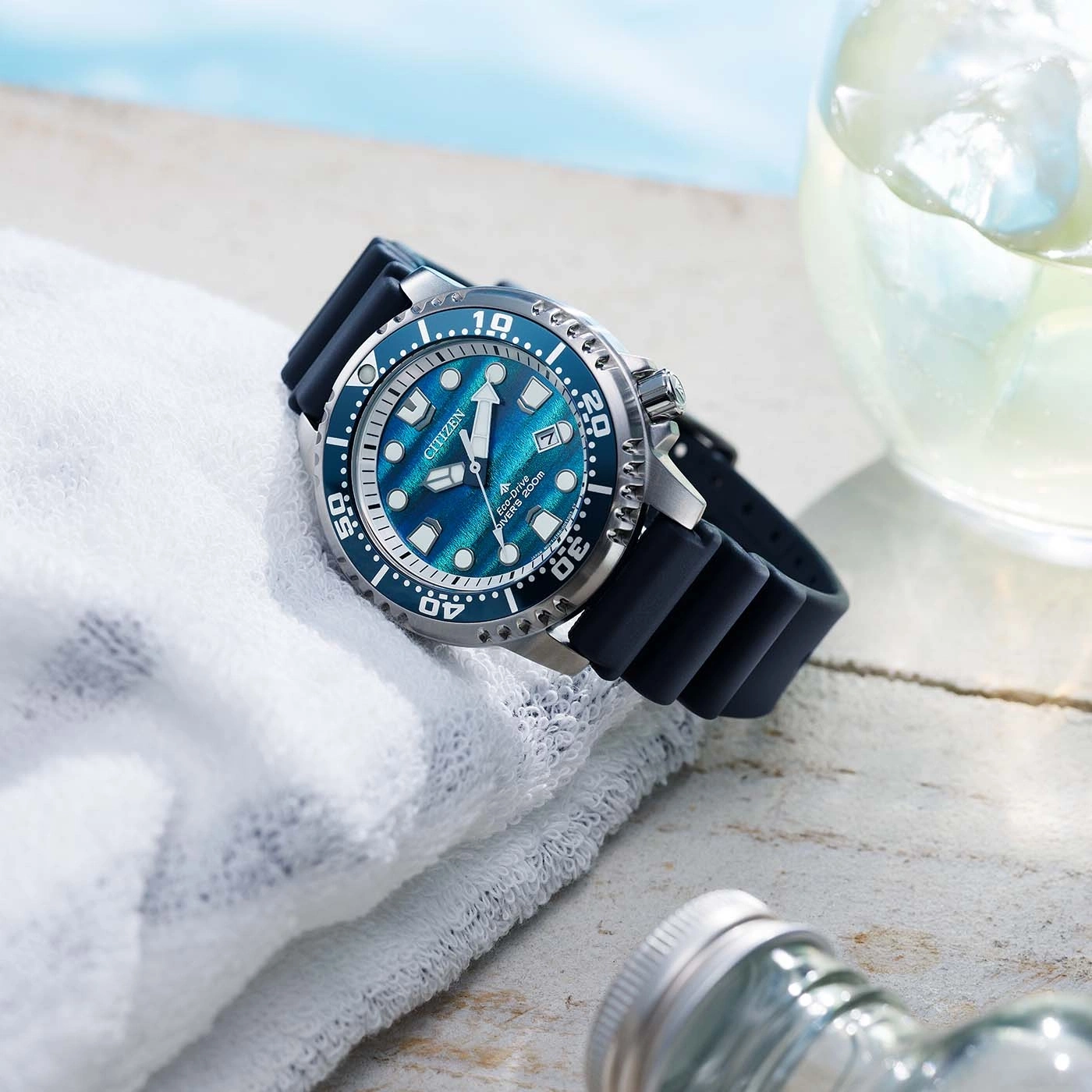 A blue Citizen Eco-Drive dive watch rests on a white towel next to a glass of iced drink.