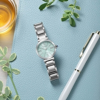 A mint green Citizen Eco-Drive watch with a silver band rests on a blue textured surface next to a white pen and a glass of amber liquid.