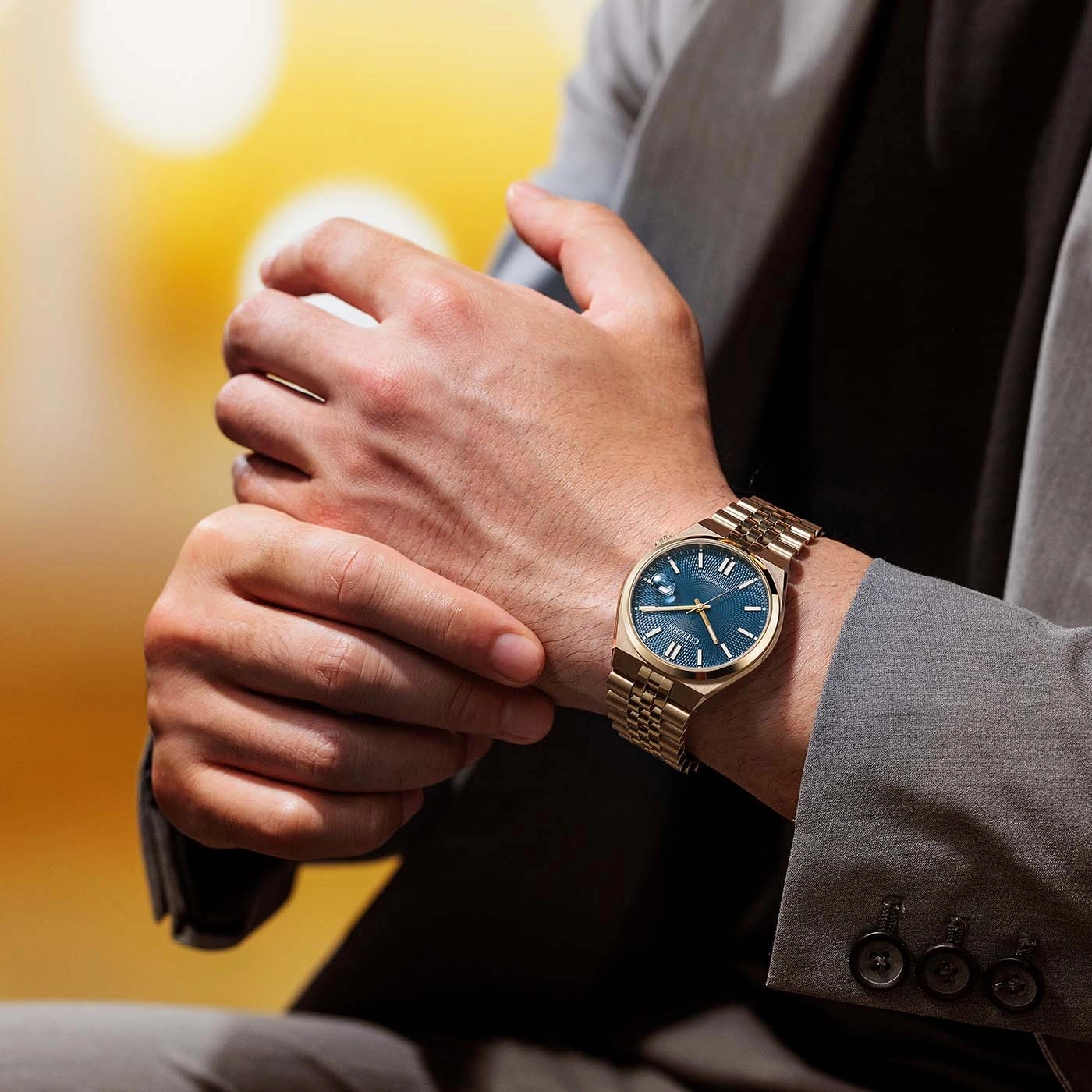 Man wearing a gold watch with a blue face and a grey suit.