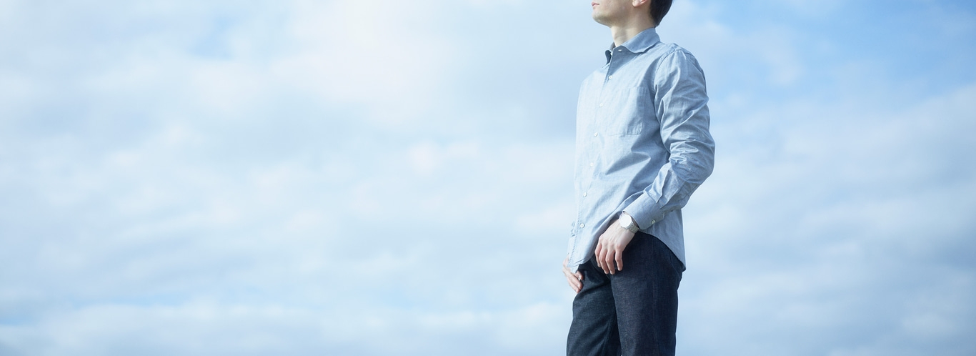 Man in shirt and jeans stands against a cloudy sky, looking up.