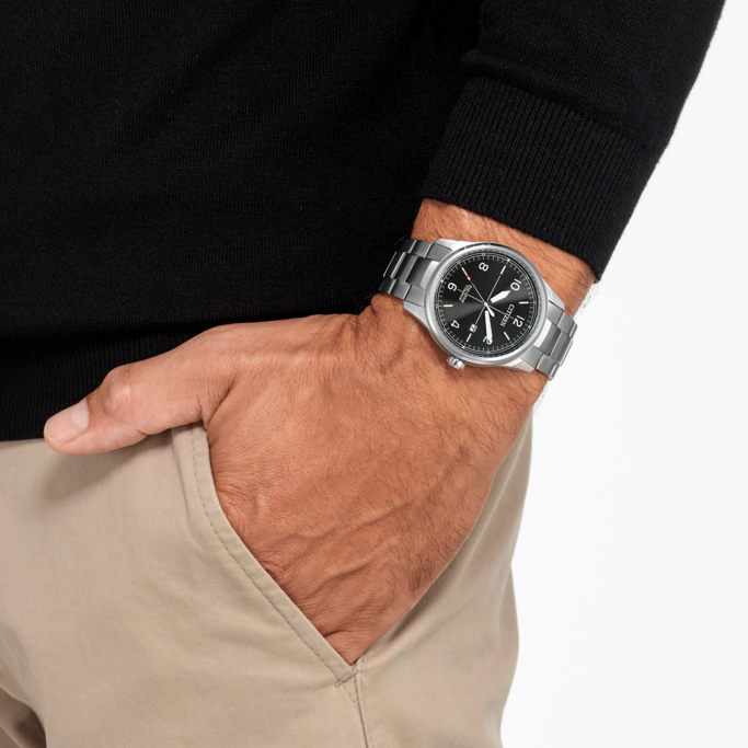Man wearing a watch, hand in pocket, black sweater, beige pants, white background.