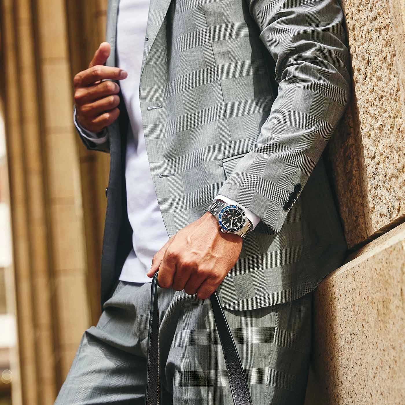 Man in suit with watch and bag leaning against a wall