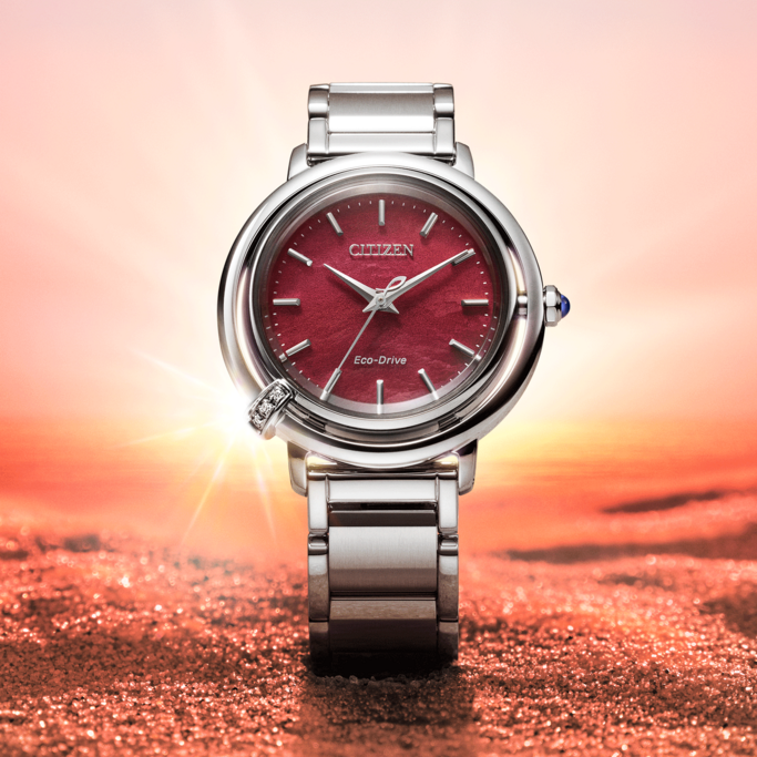 A silver Citizen Eco-Drive watch with a red face is prominently displayed against a shimmering red and orange background, evoking a sunset or desert scene.
