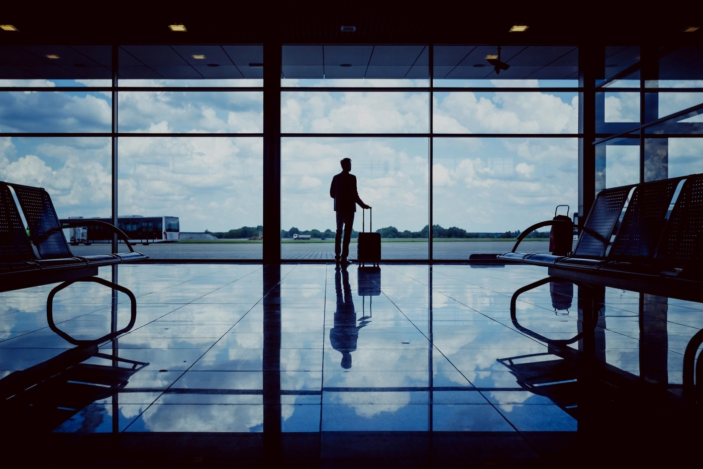 Silhouette of a person with luggage in an airport, looking out the window.