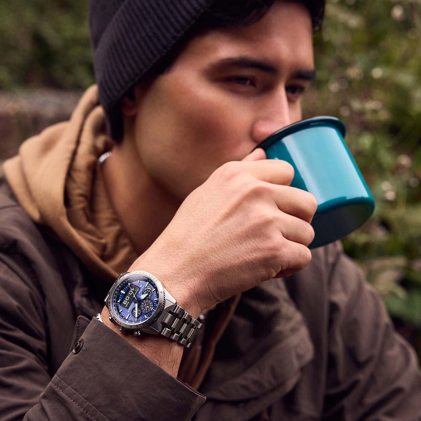 Man drinking from a mug, wearing a watch and a beanie in nature.