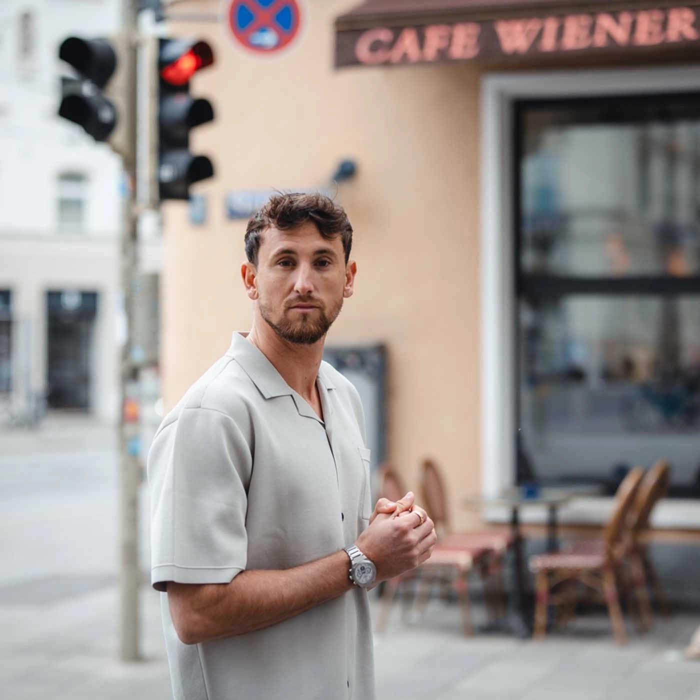Man in front of a cafe, wearing a light shirt and a watch, looking at the camera.