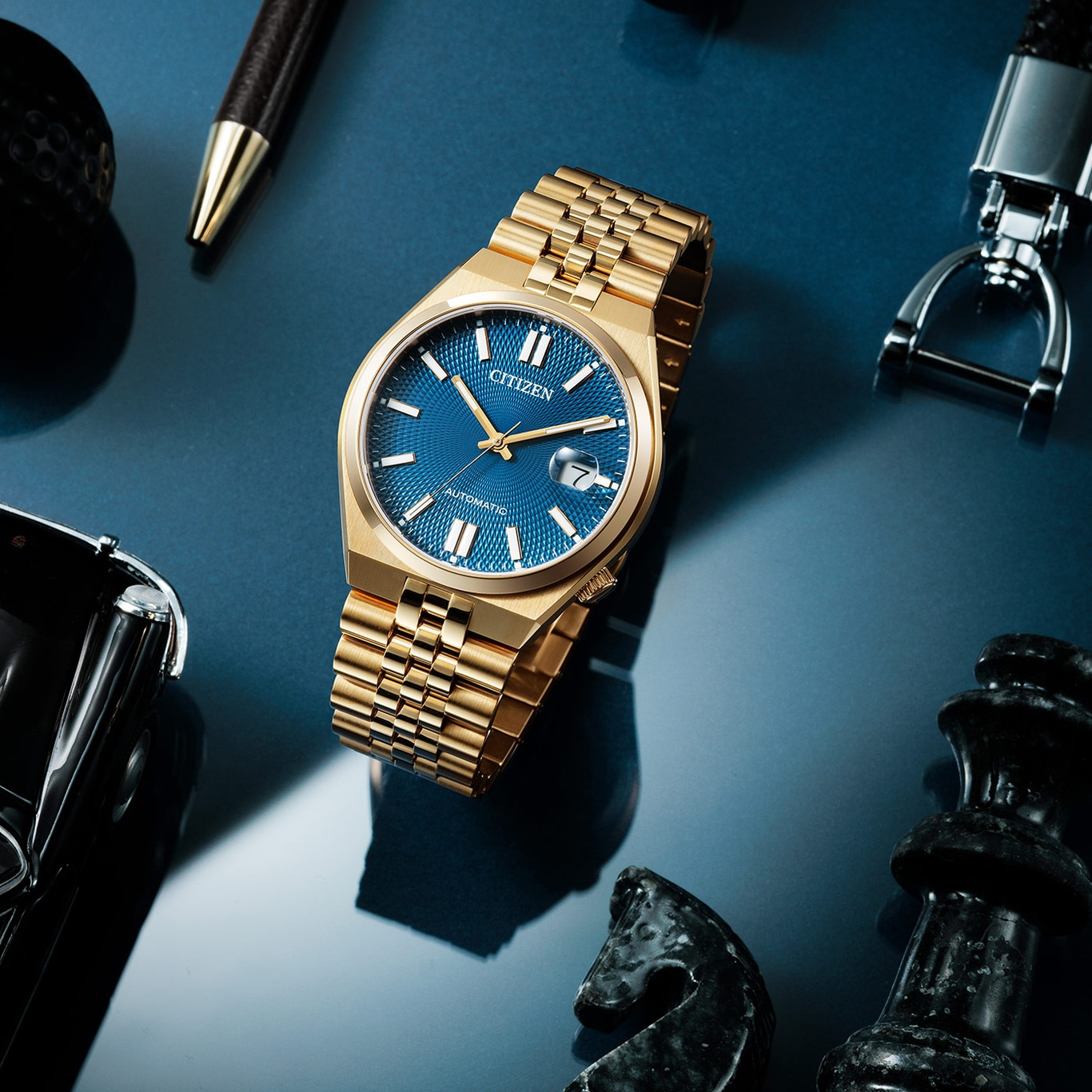 Gold Citizen watch with blue face, pen, chess pieces, and keychain on blue surface.