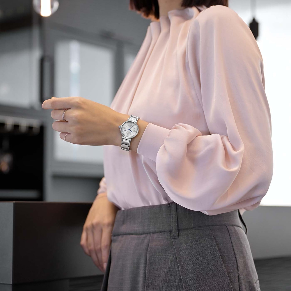 Woman wearing a pink blouse and grey pants, checking her watch.
