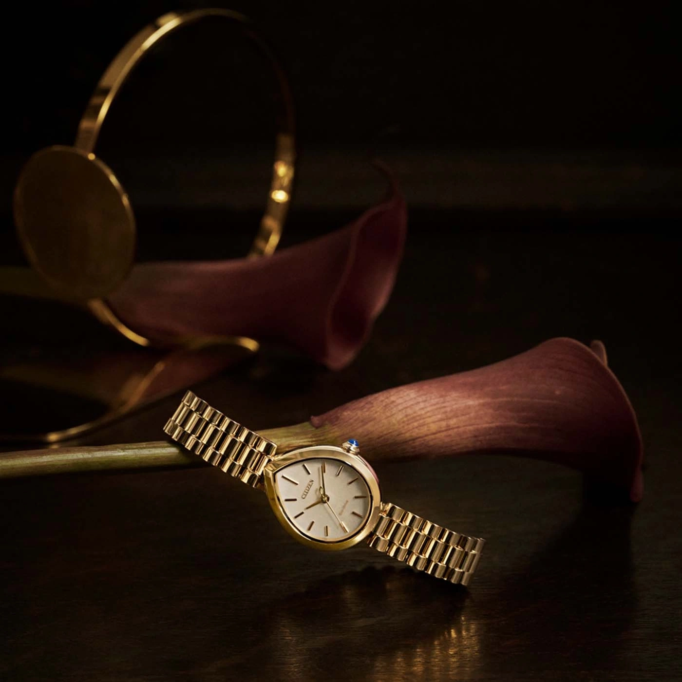 A gold watch with a bracelet rests on a dark surface with flowers.