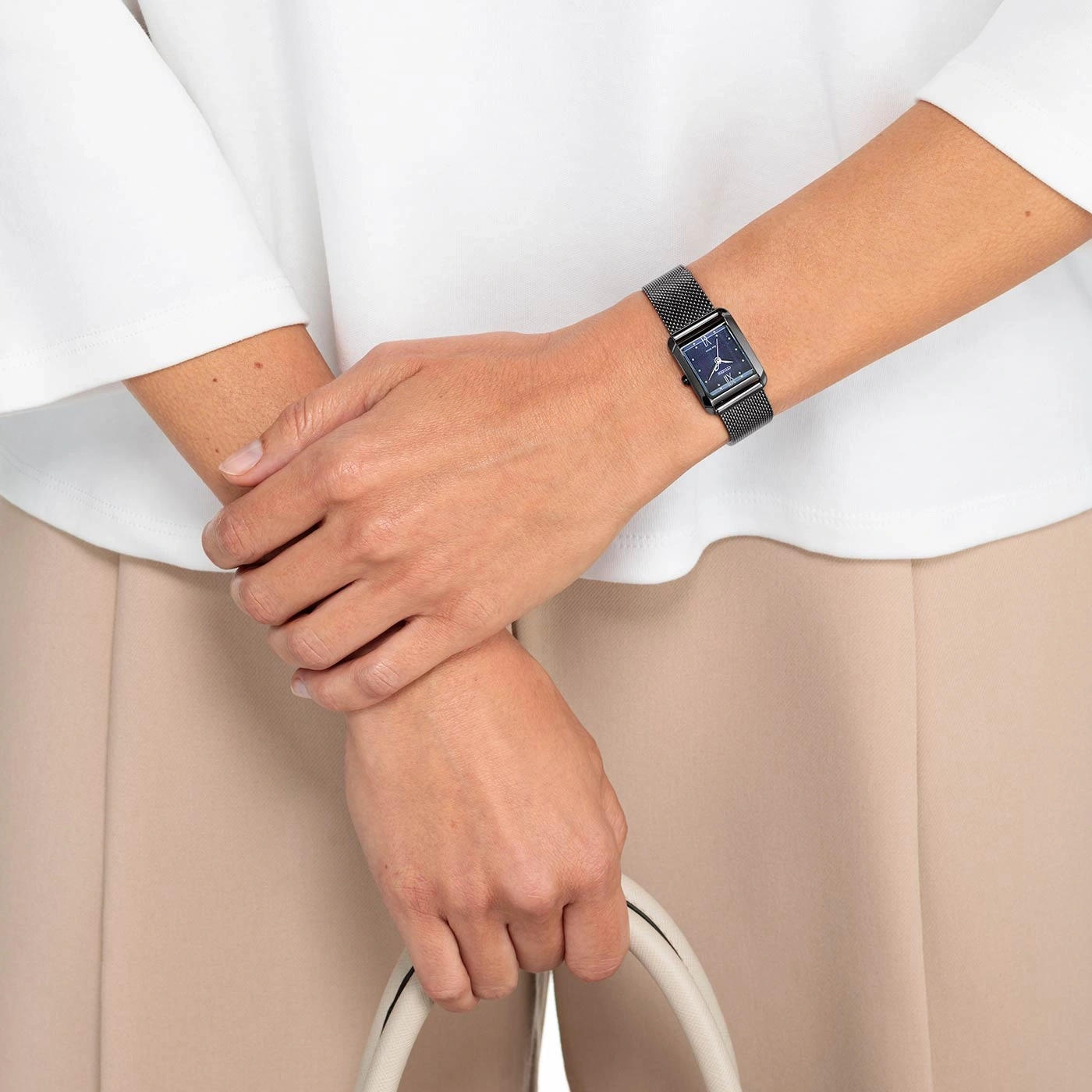 Woman wearing a black watch, holding a white bag, beige pants, white shirt.