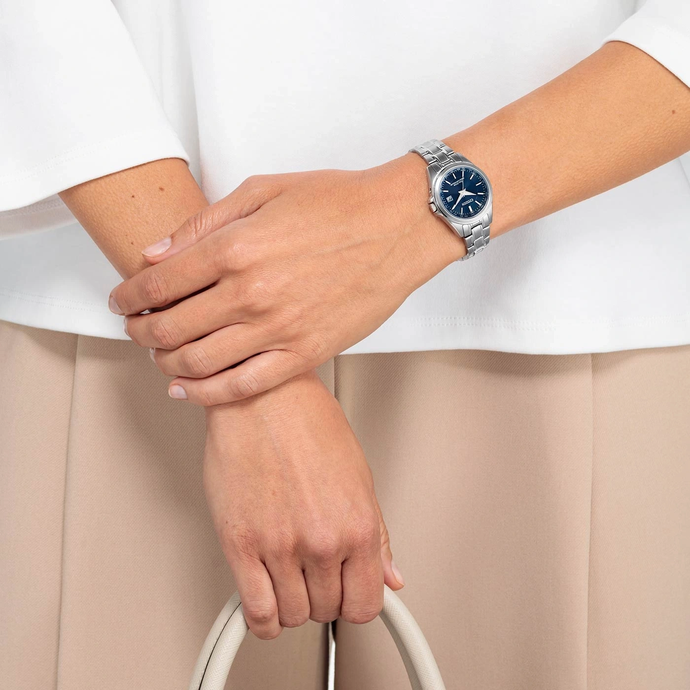 Woman wearing a watch, holding a bag, with white shirt and beige pants.
