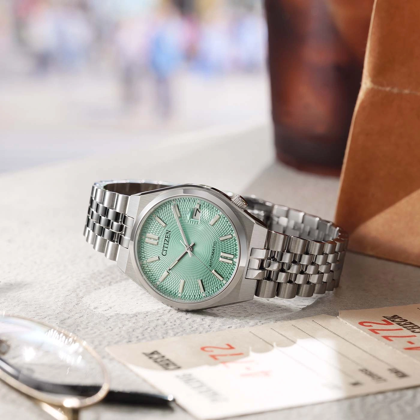 A Citizen automatic watch with a mint green dial rests on a textured surface near glasses and papers.