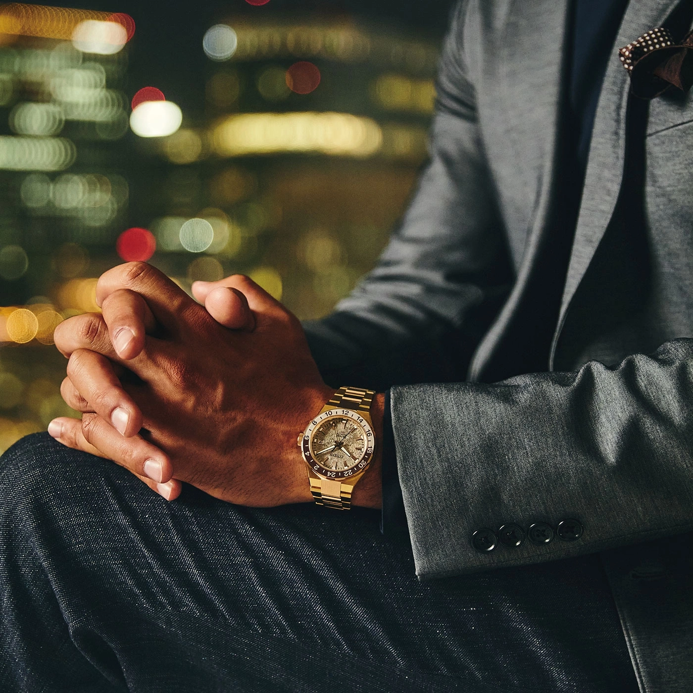 Man in suit with gold watch, hands clasped, city lights in background.