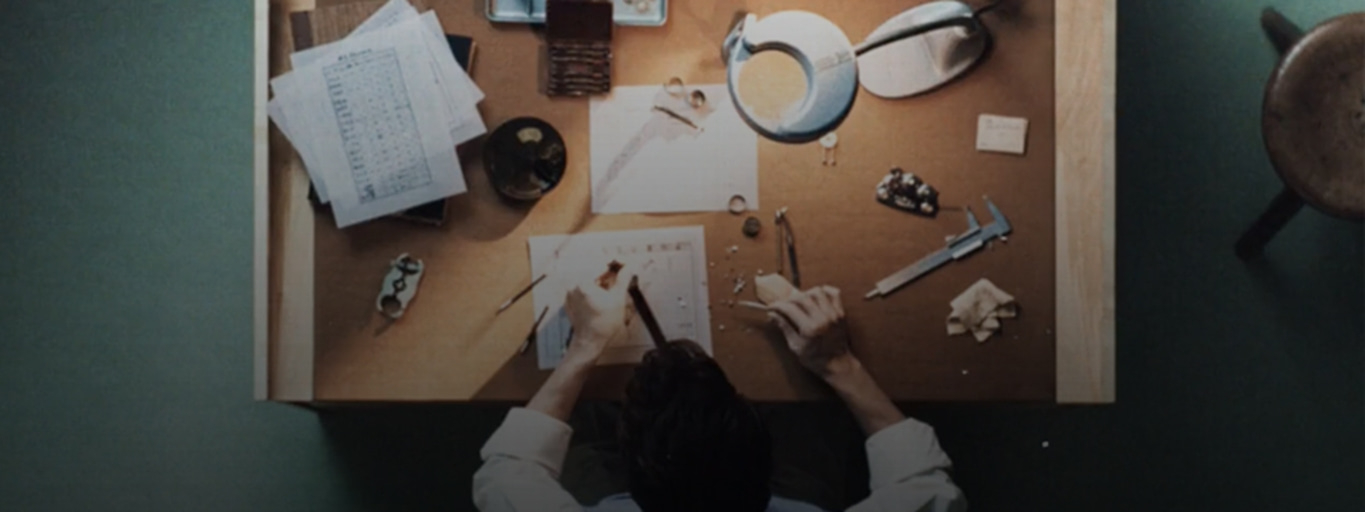 Jeweler at work, crafting jewelry on a wooden desk with tools and papers.