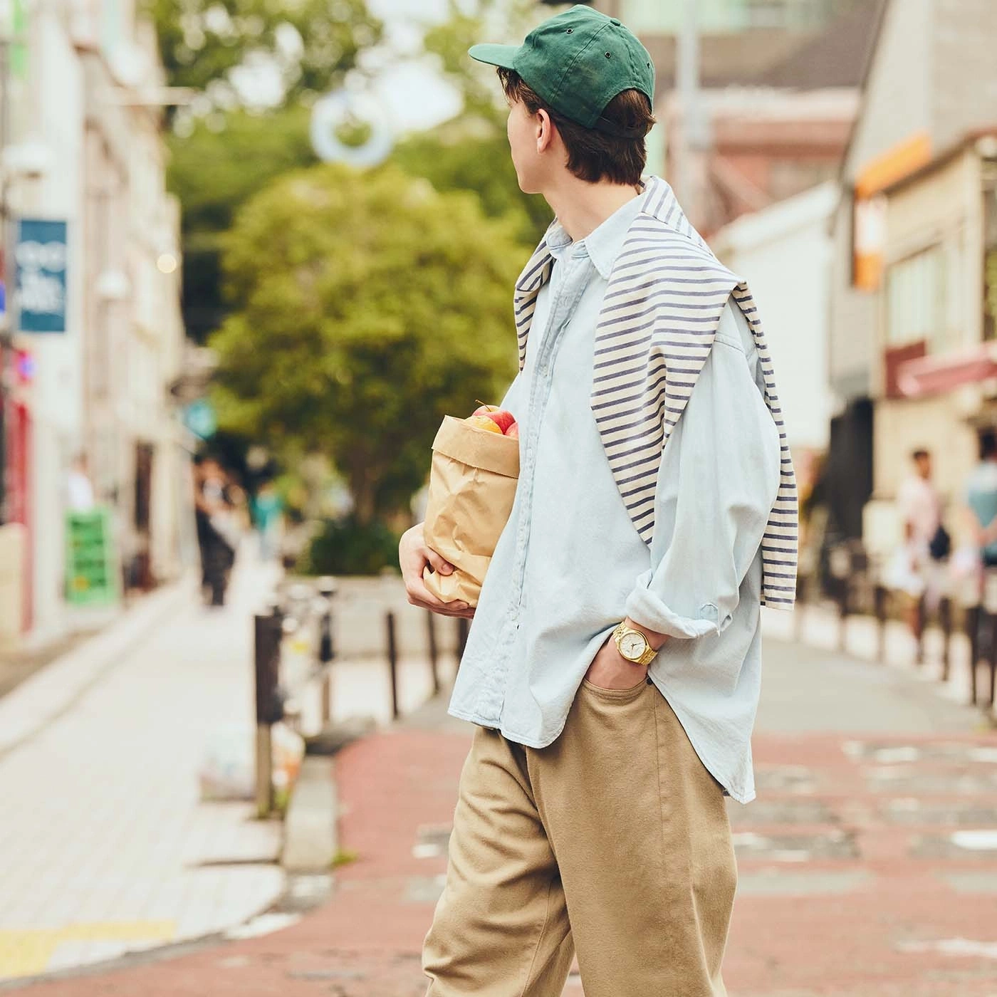 A man in a cap and light blue shirt walks down a street holding groceries.