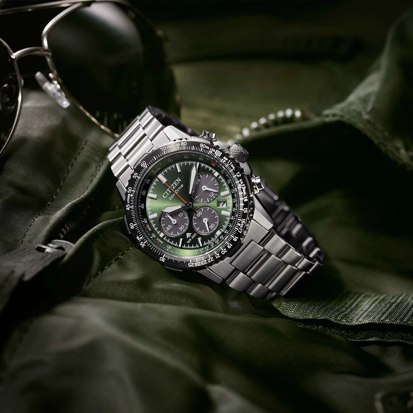 Citizen watch with green face and metal band on green fabric with sunglasses.