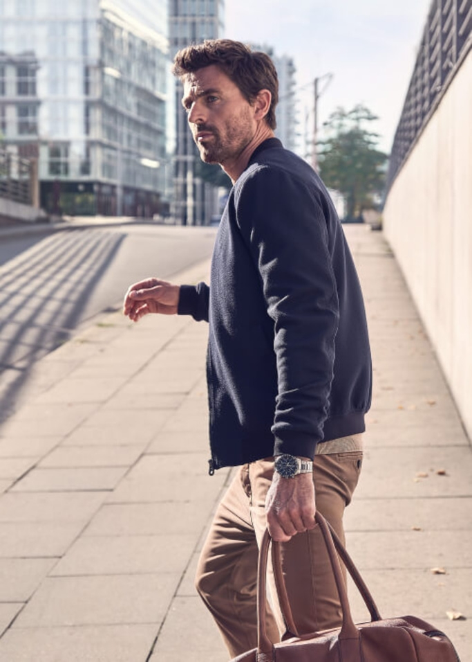 Man walking on sidewalk, holding a bag, wearing a jacket and watch.