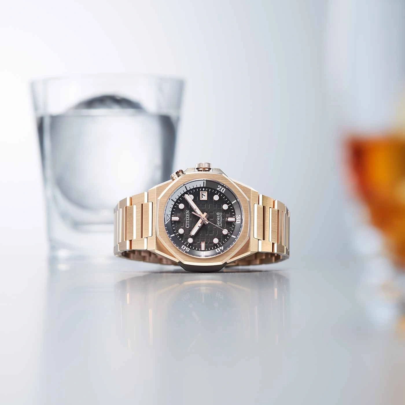 Gold watch with black face, glass of water, and reflective surface.