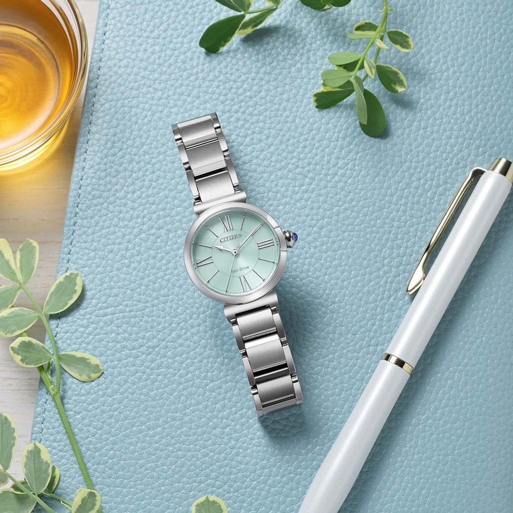 A mint green Citizen Eco-Drive watch with a silver band rests on a blue textured surface next to a white pen and a glass of amber liquid.