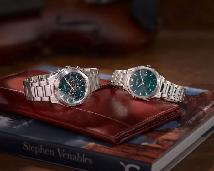 Two Citizen watches with green faces on a book and leather wallet.