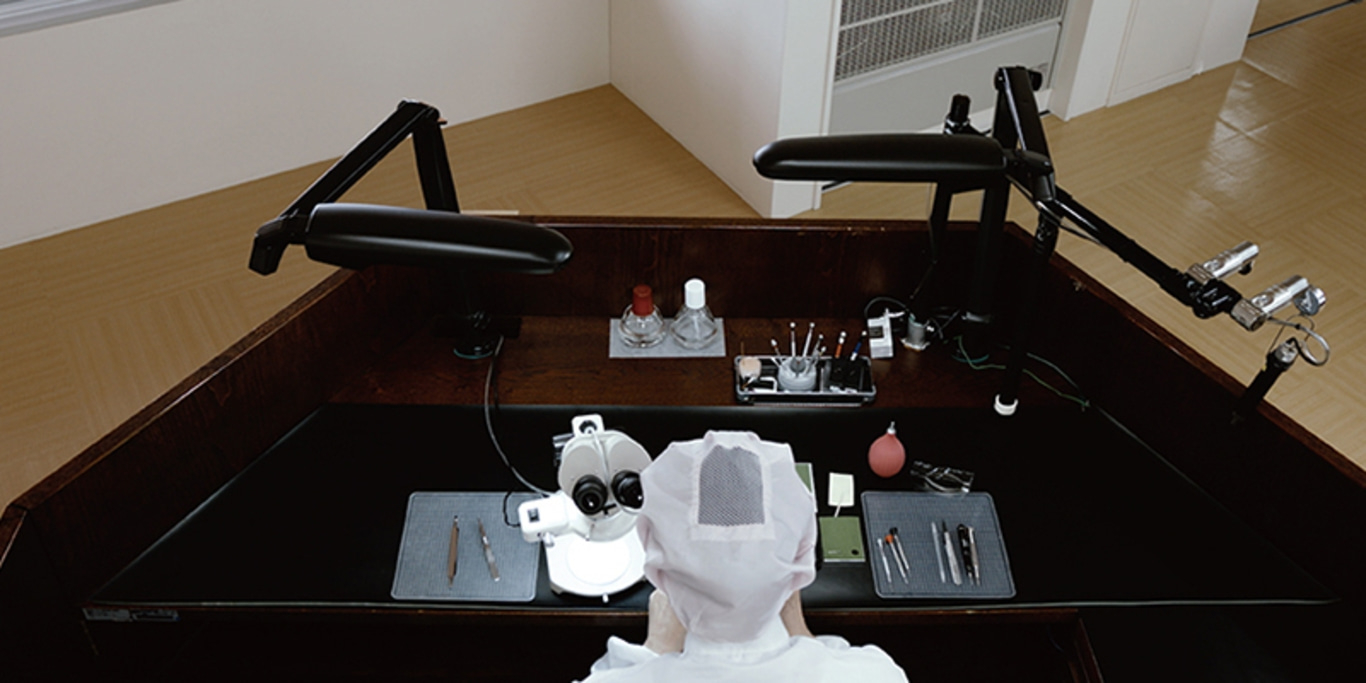 A person in a lab coat works at a desk with a microscope and tools.
