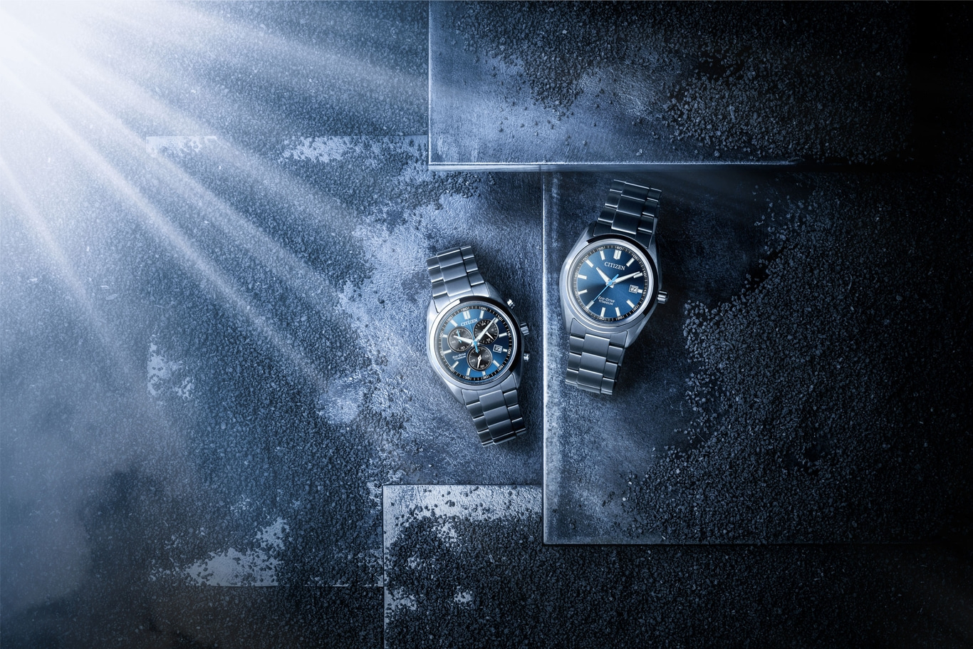 Two Citizen Eco-Drive titanium watches on a dark textured surface with light rays.
