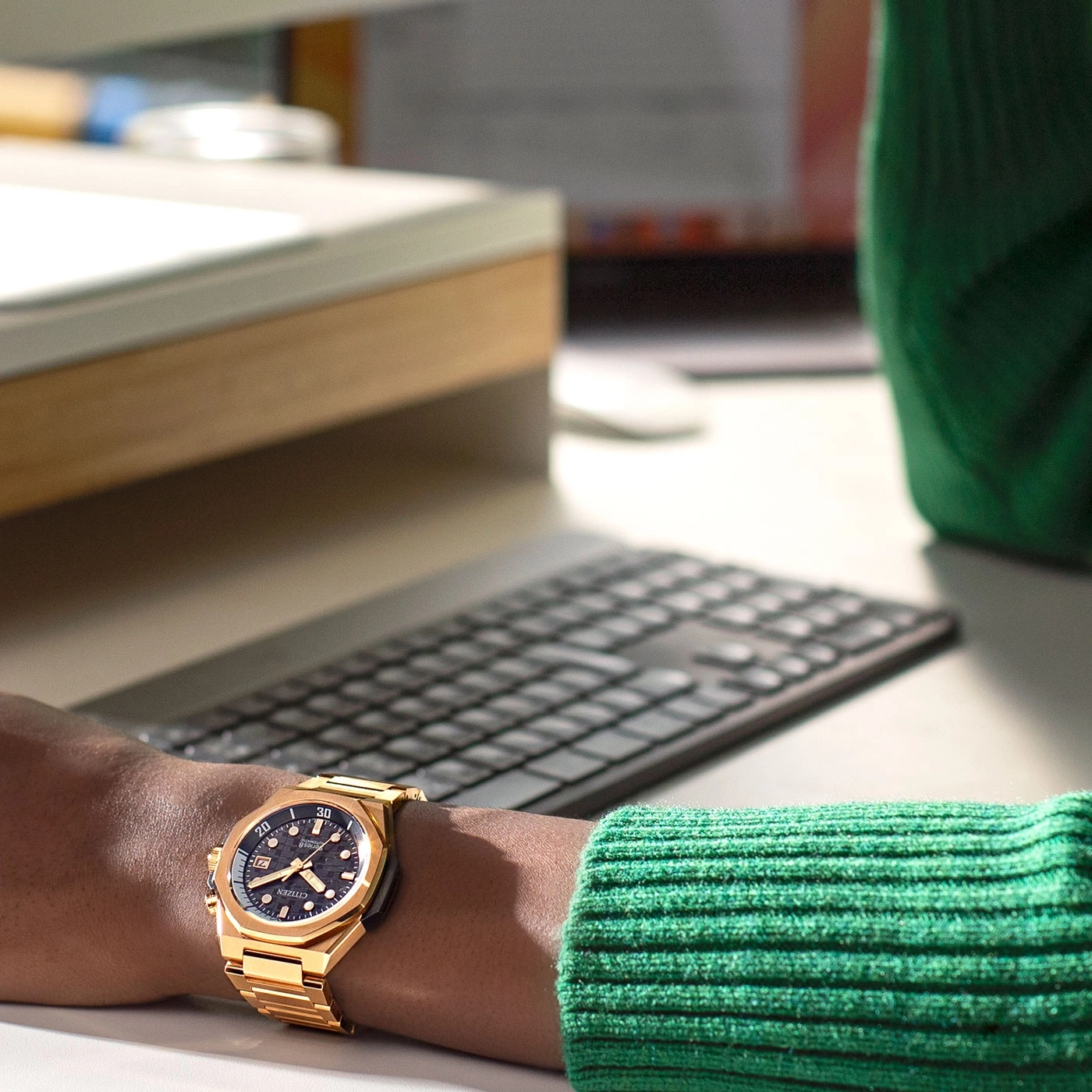 Gold watch on wrist, green sweater, keyboard, desk, office setting, Citizen watch