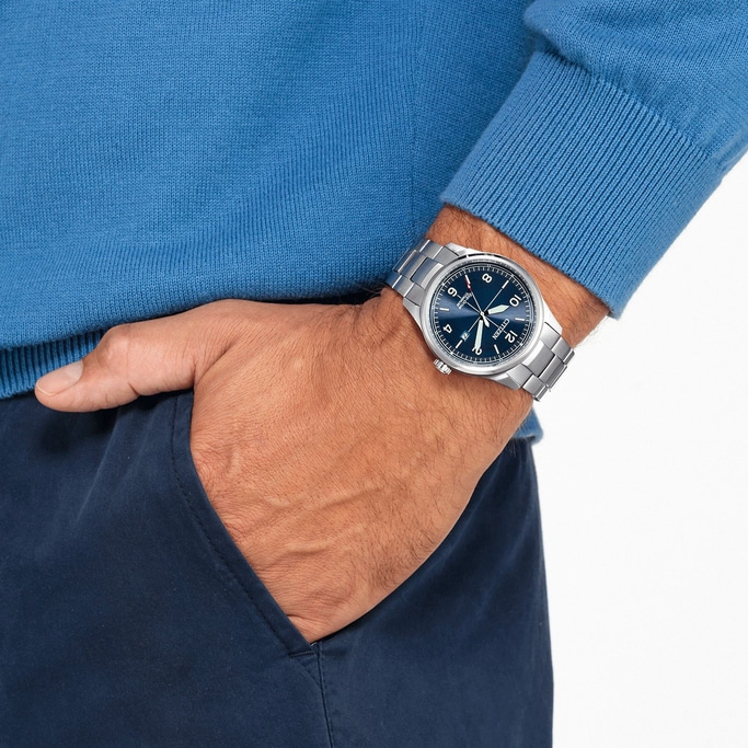 Man wearing a watch, blue sweater, and navy pants with hand in pocket.