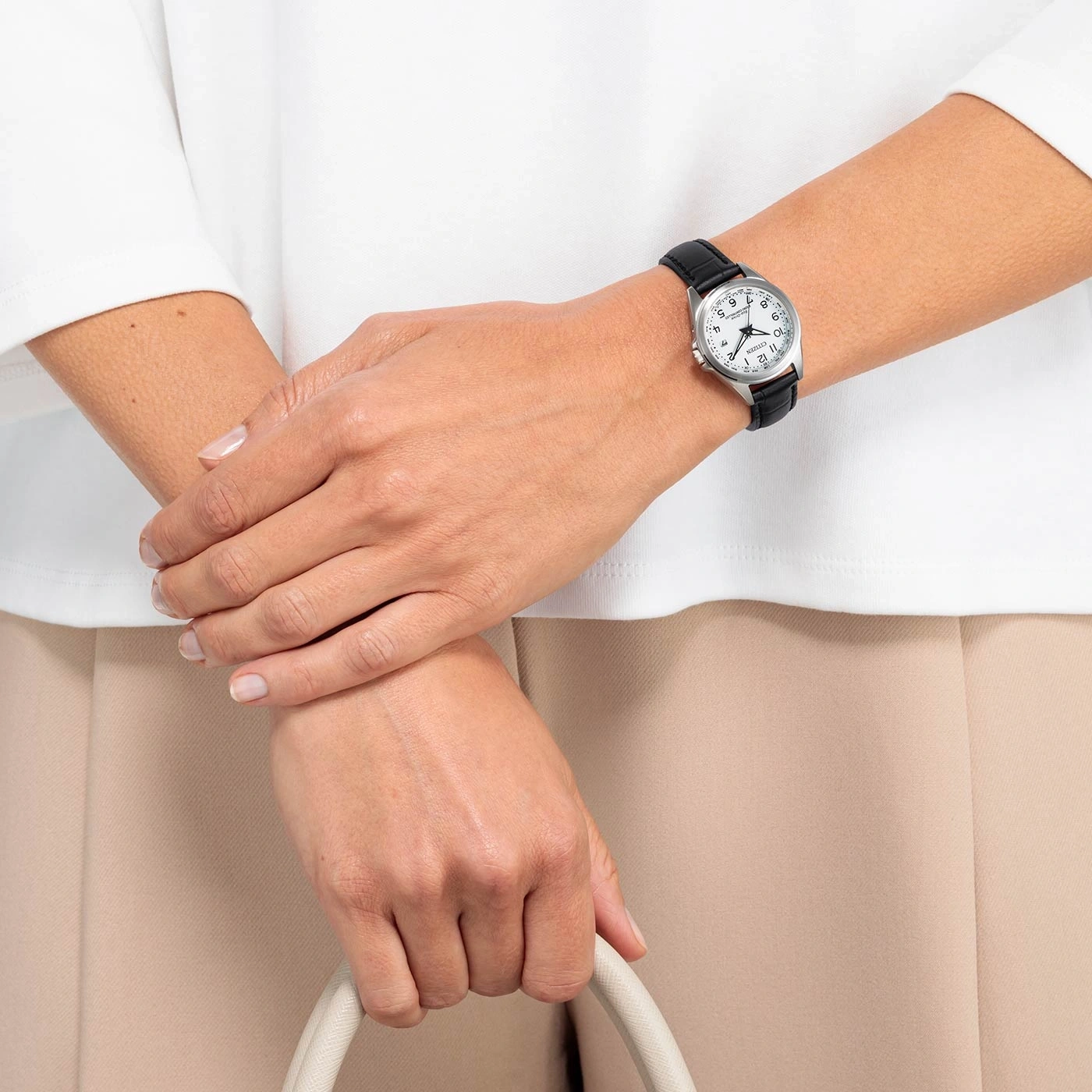 Woman wearing a watch, holding a bag, with white shirt and beige pants.