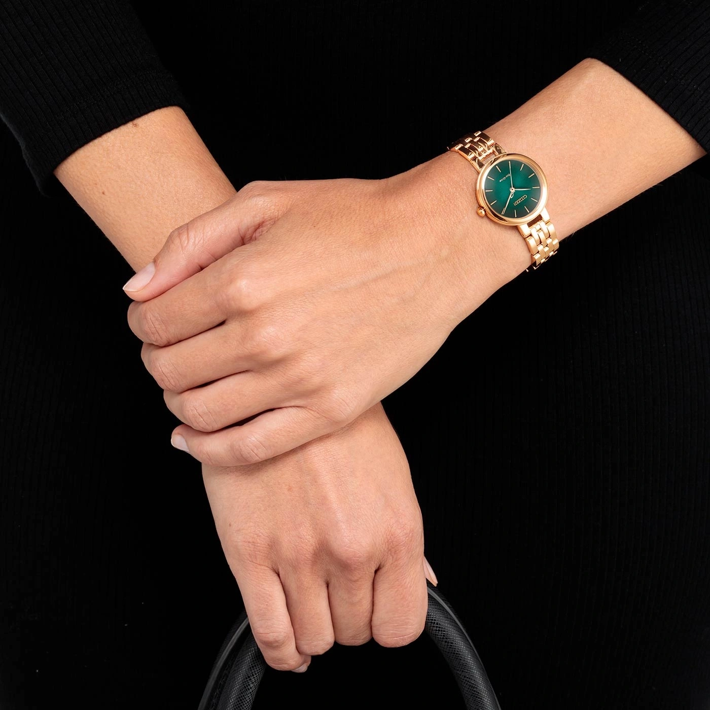 Woman wearing a gold watch with a green face, black dress and a black bag.
