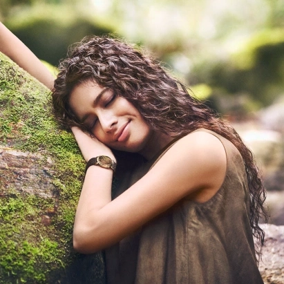 A woman with curly hair rests her head on a mossy tree trunk, eyes closed and a gentle smile on her face, set against a blurred natural background.

