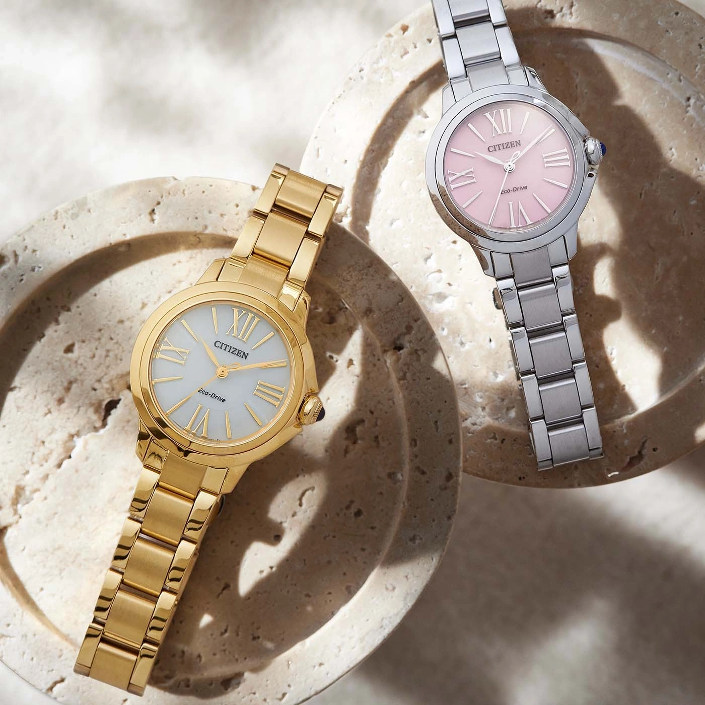 Two Citizen watches, gold and silver, on a textured surface.