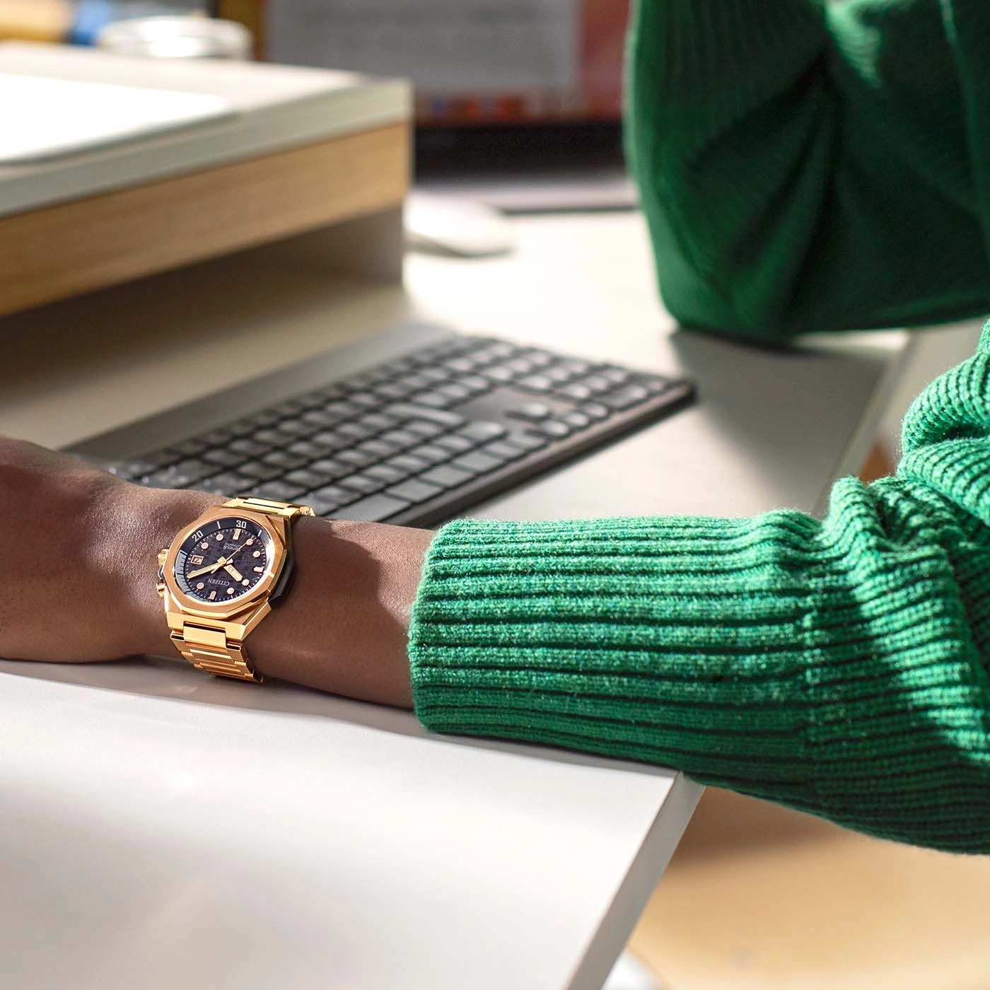 Gold watch on wrist, green sweater, keyboard, white desk, office setting.