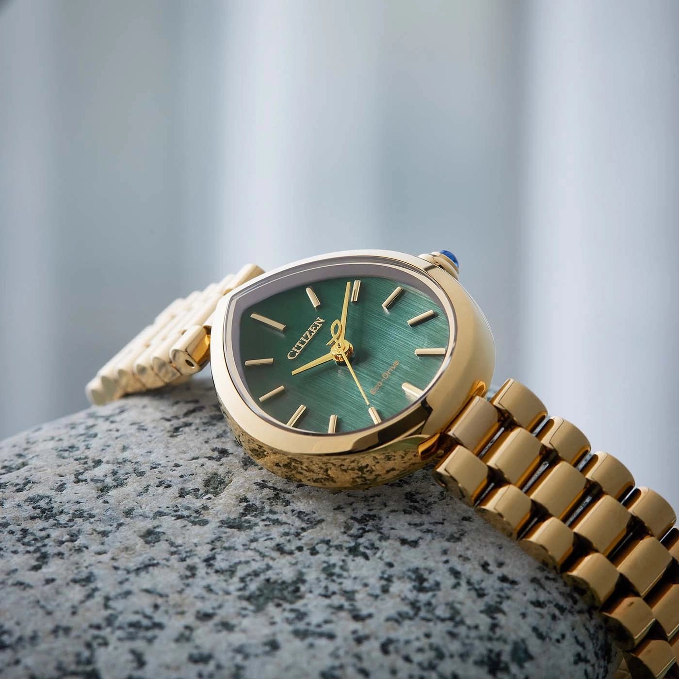 A gold Citizen Eco-Drive watch with a green face rests on a rock.