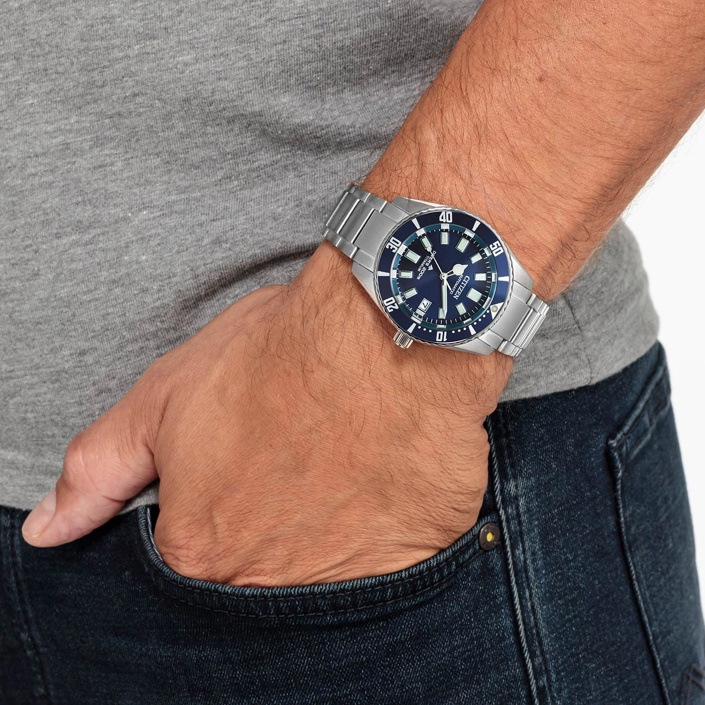 Man wearing a Citizen watch, blue dial, stainless steel bracelet, jeans and grey shirt.