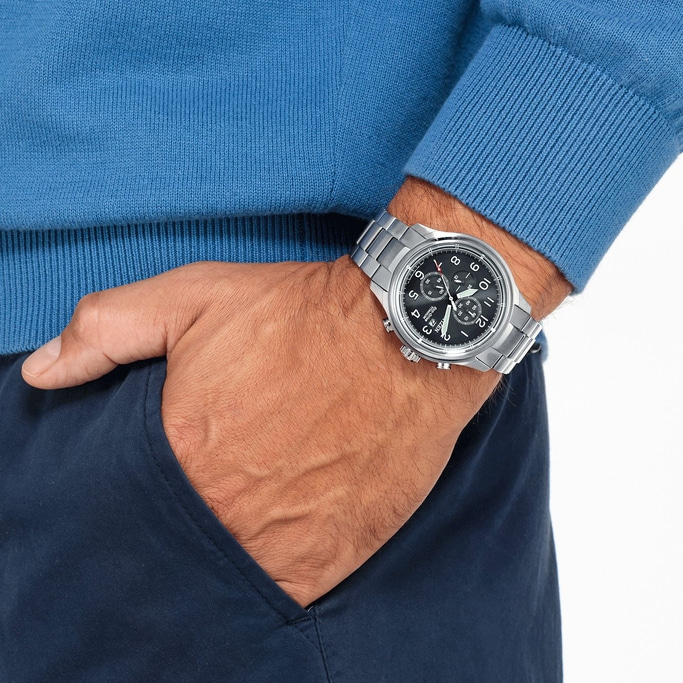Man wearing a watch, blue sweater, and navy pants with hand in pocket.
