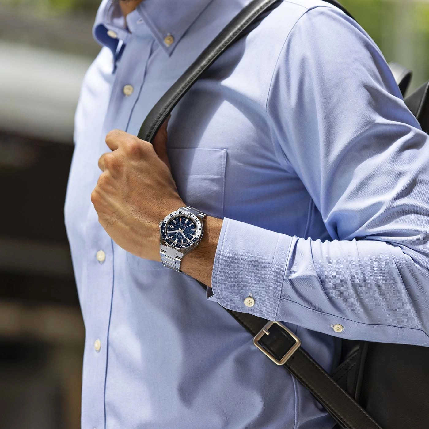 Man wearing a watch and blue shirt with a black bag on his shoulder.