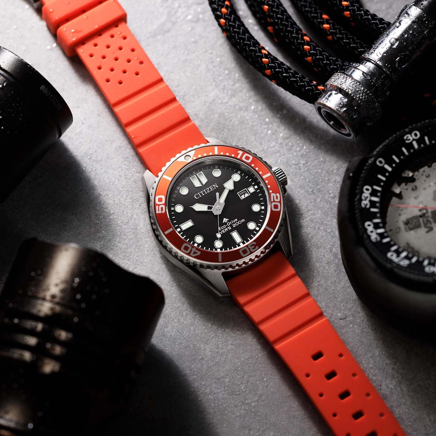 A close-up shot of a Citizen Eco-Drive diver's watch with a red strap, next to a compass and flashlight.