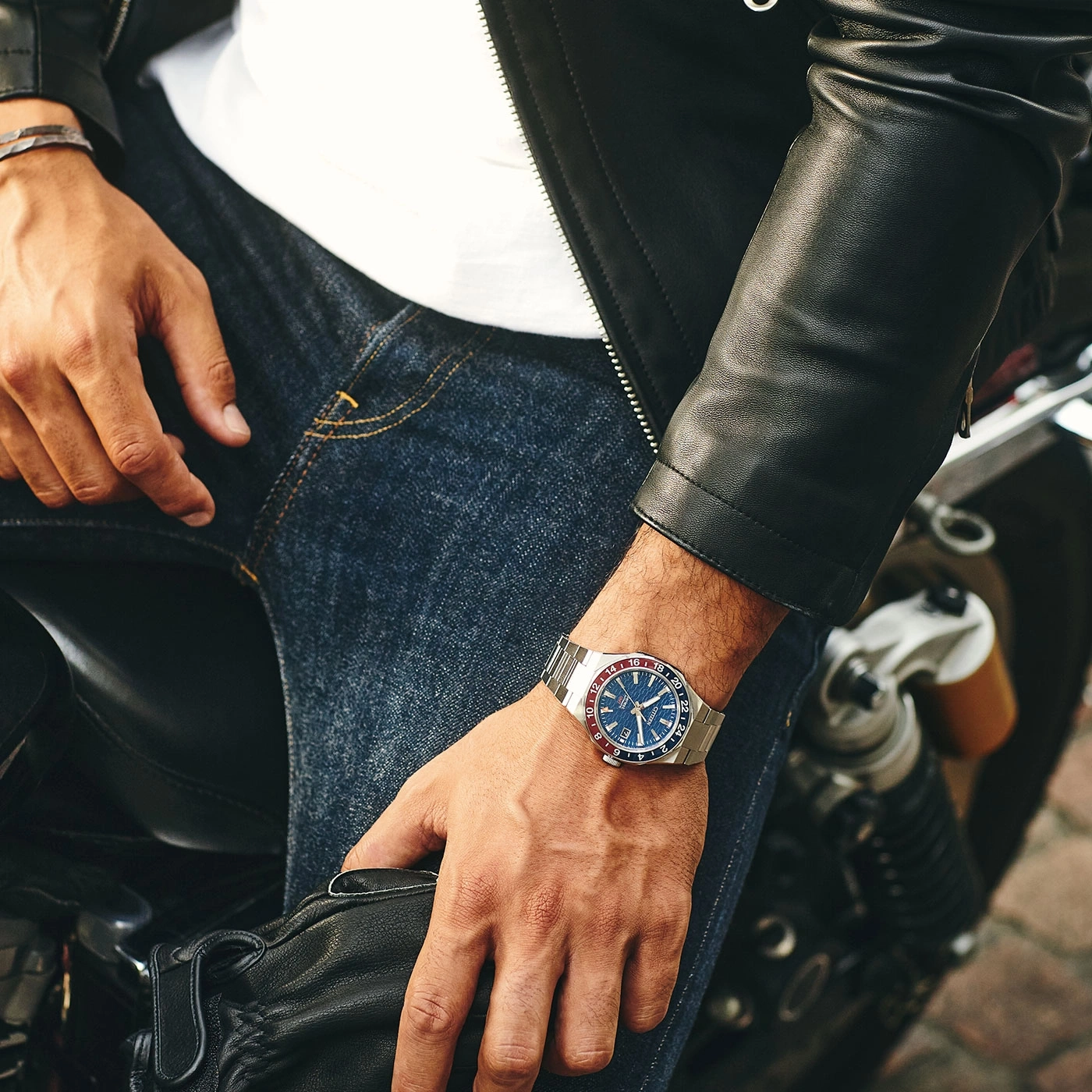 Man wearing a watch, leather jacket, and jeans on a motorcycle.