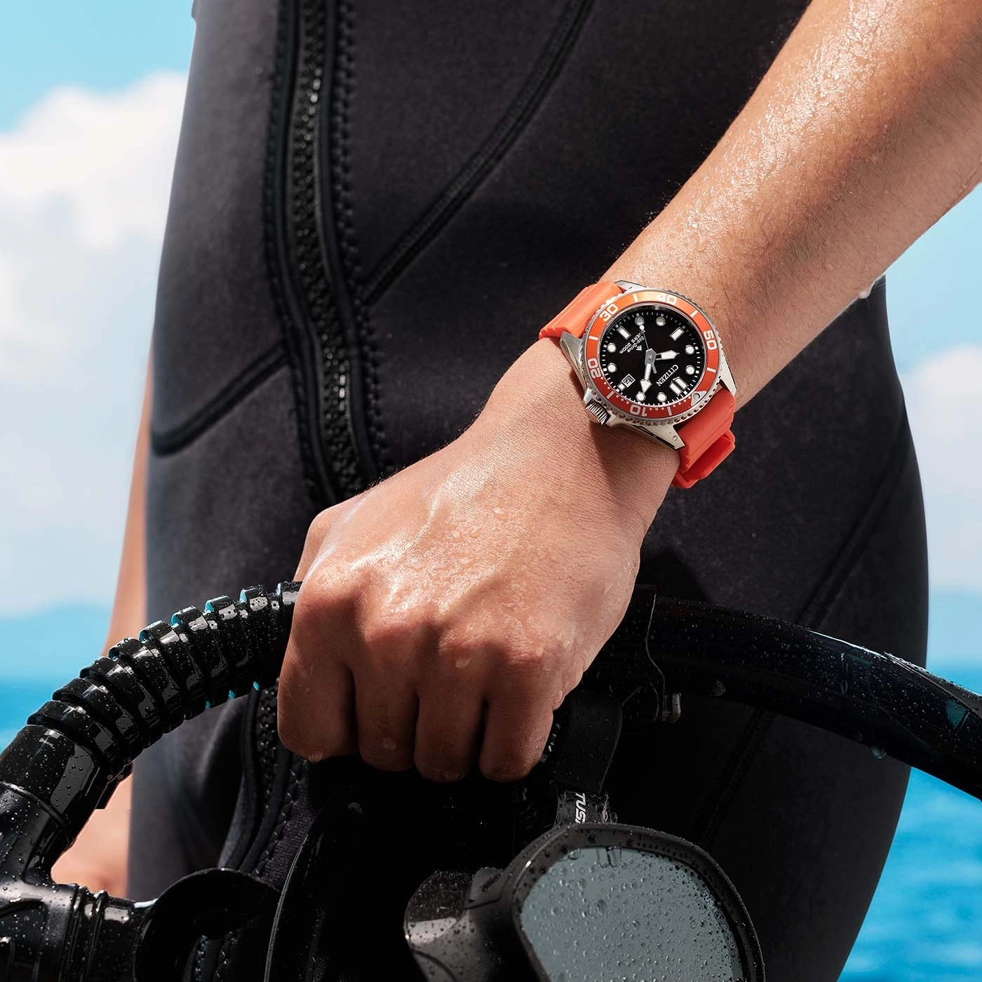 A person in a wetsuit holds diving gear, wearing a Citizen watch.