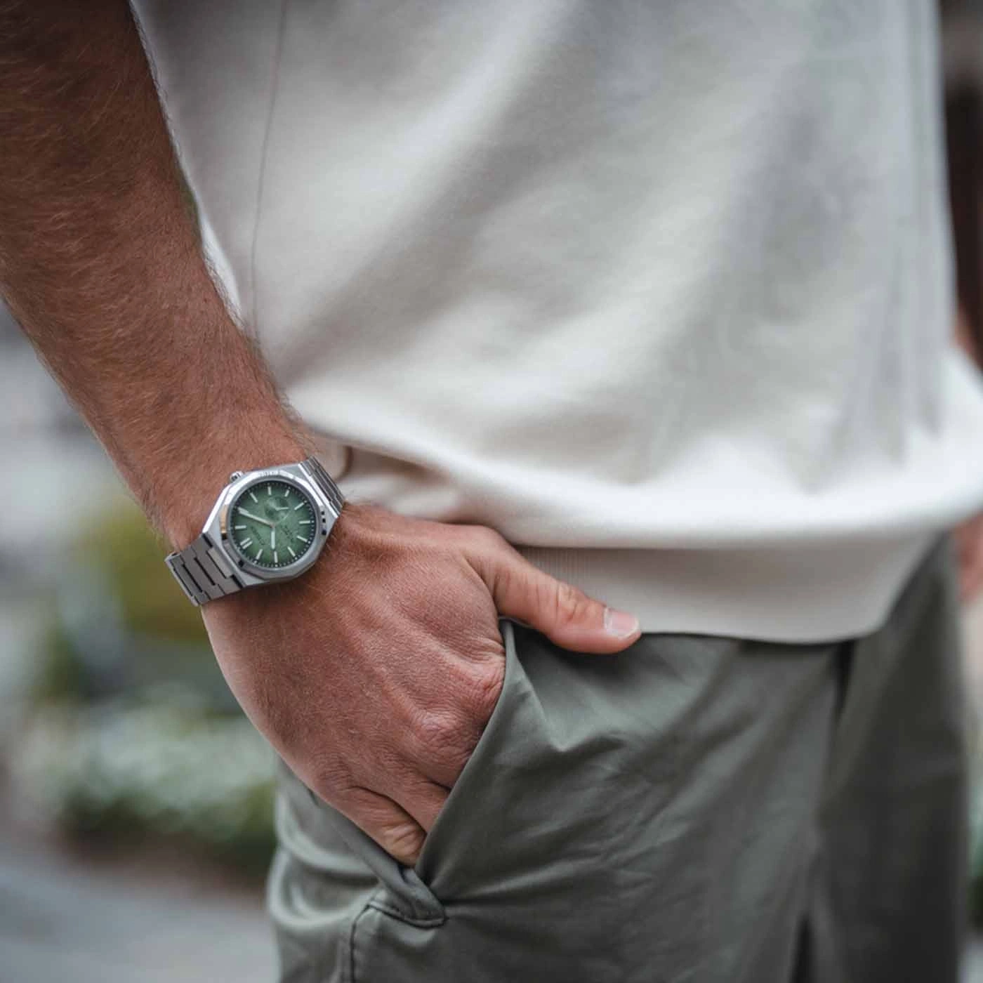 Man wearing a silver watch with a green face, hand in pocket.