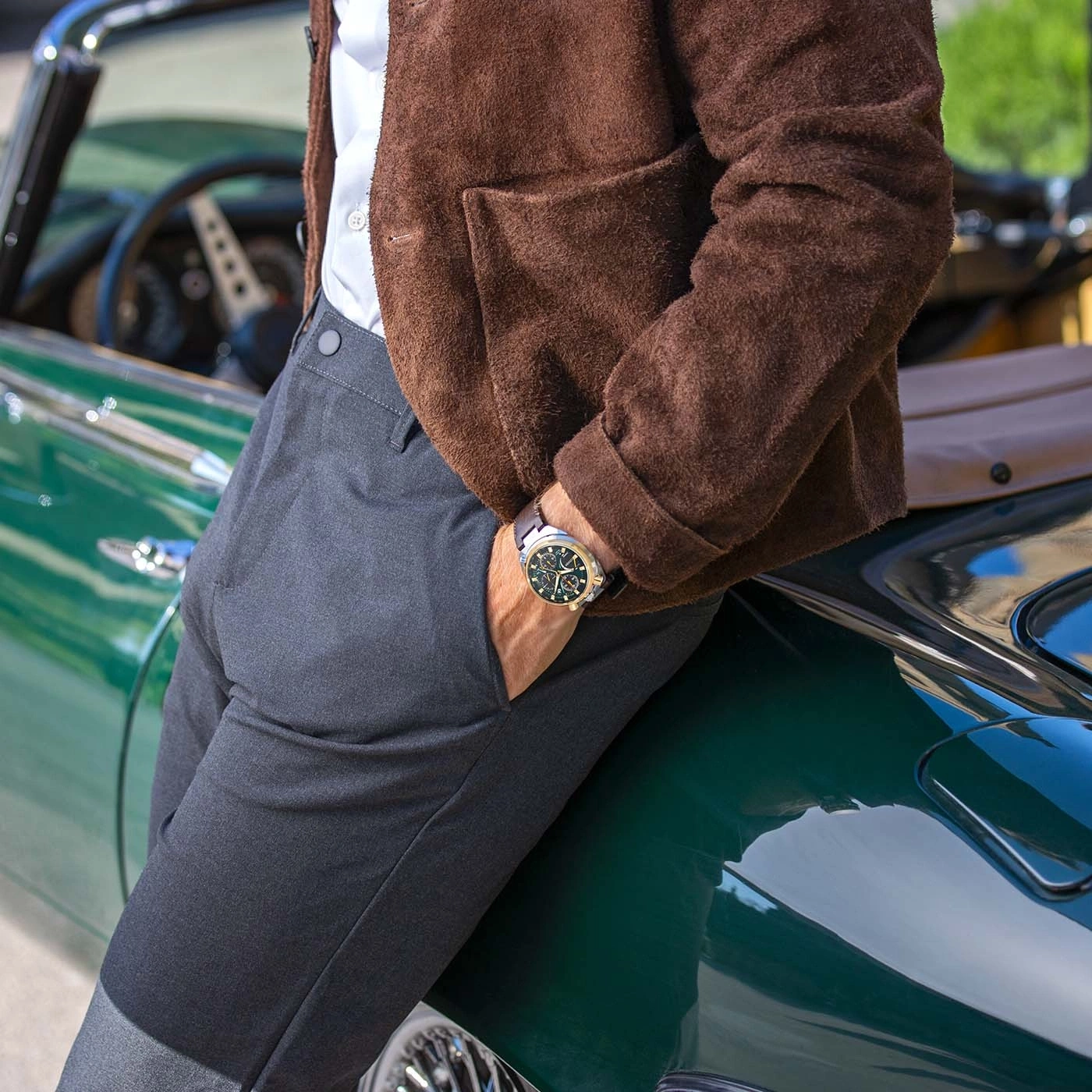 Man leaning against a vintage green car, wearing a brown jacket and a watch.