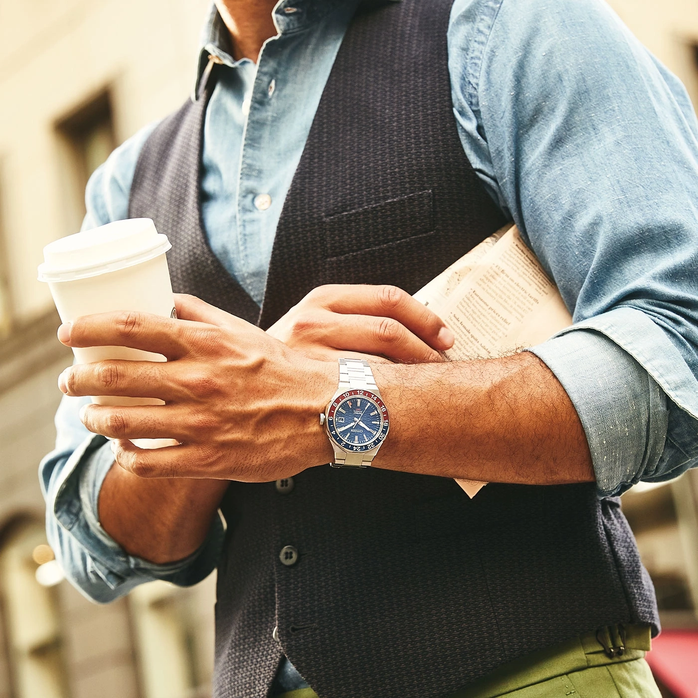 Man holding coffee and newspaper, wearing a watch and vest, urban setting.